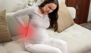 Pregnant,Woman,Sitting,On,Couch,With,Hand,On,Back,Experiencing